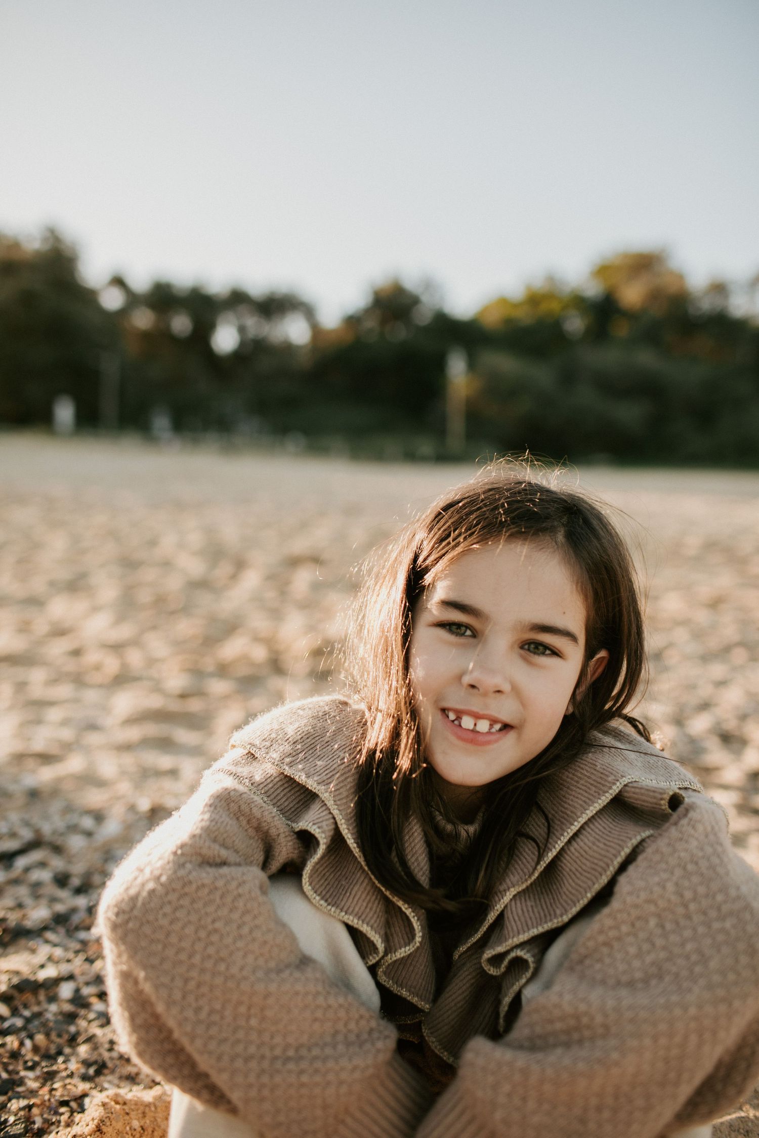 Smiling young girl with dark hair sitting on a pebble beach in a beige knit coat.