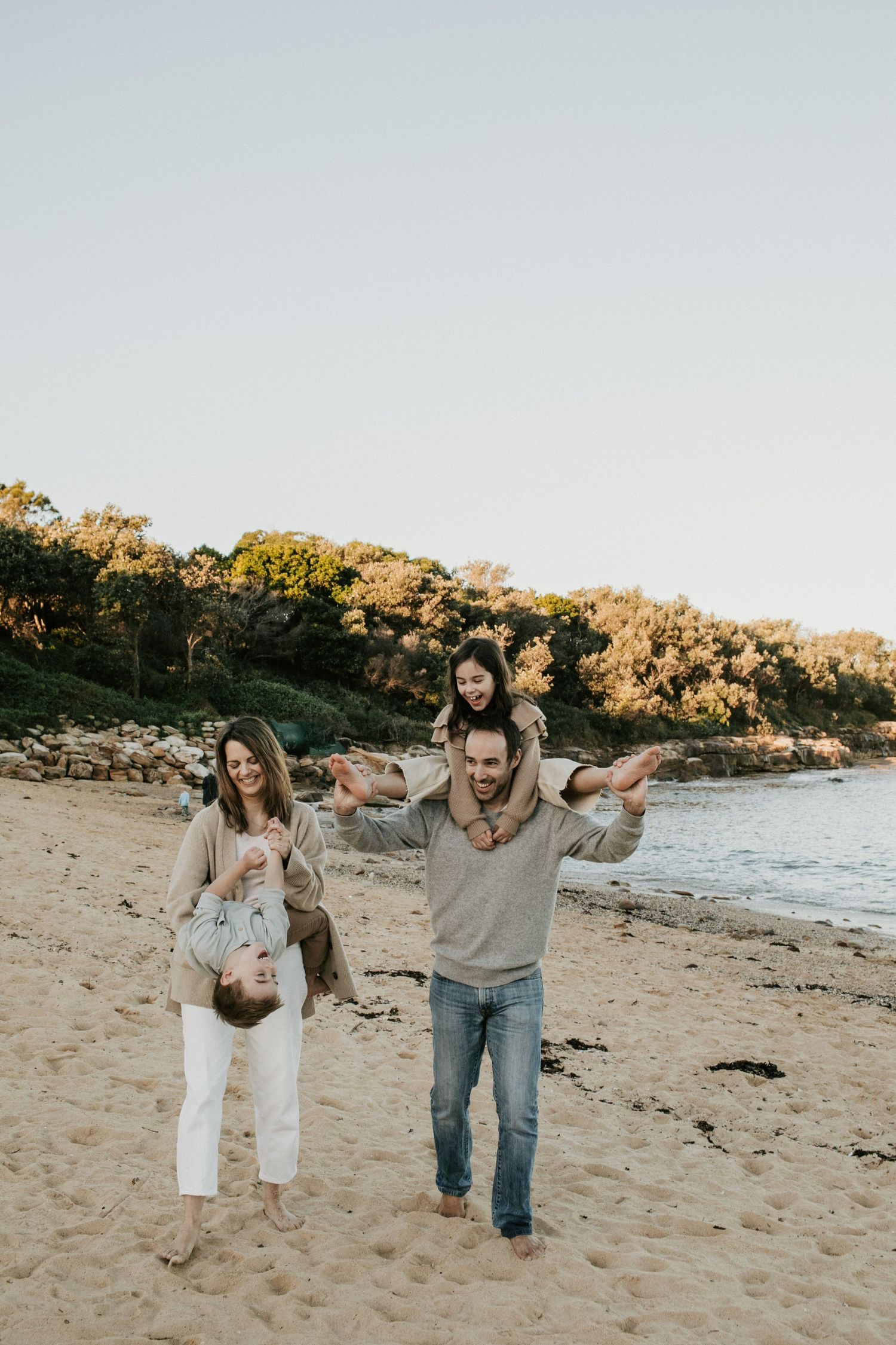 Joyful family of four playing on a sandy beach with dad carrying girl on shoulders at sunset.
