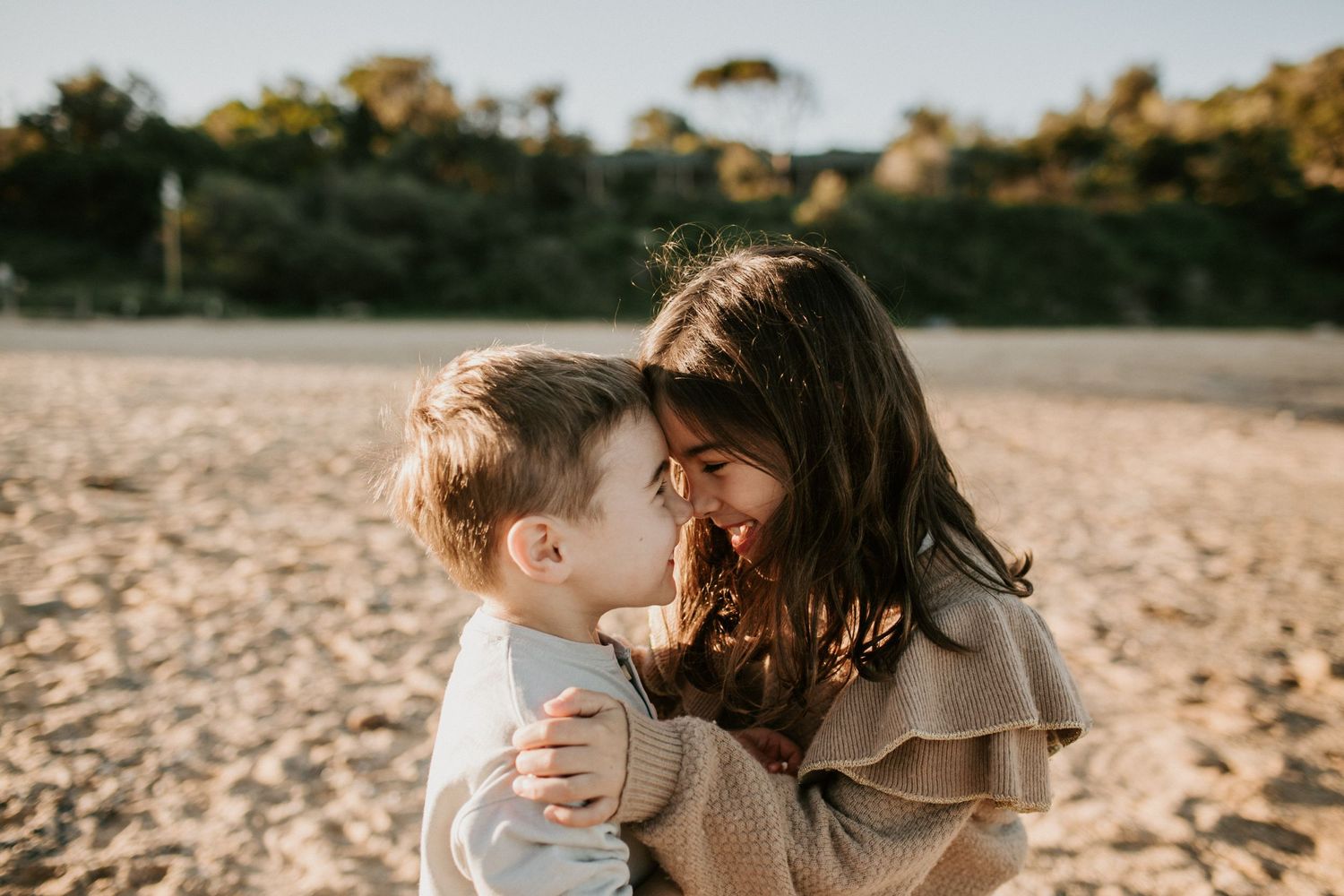 Two young siblings sharing a tender nose-to-nose moment while embracing on a sandy beach.