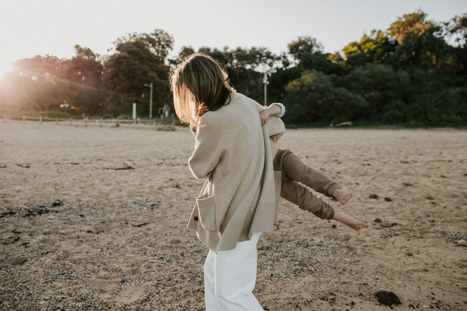 Woman in beige cardigan and white pants walking on a sandy beach in sunlight.