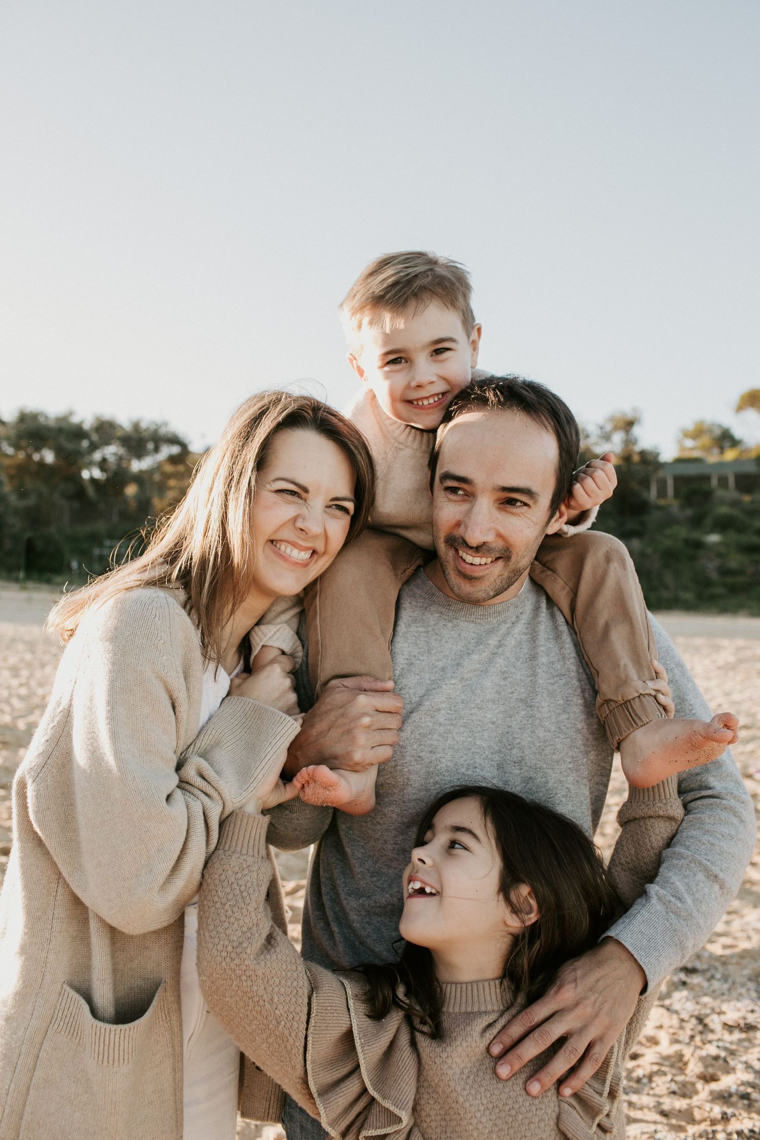 Smiling family of four poses together on sandy beach with boy on fathers shoulders at sunset.