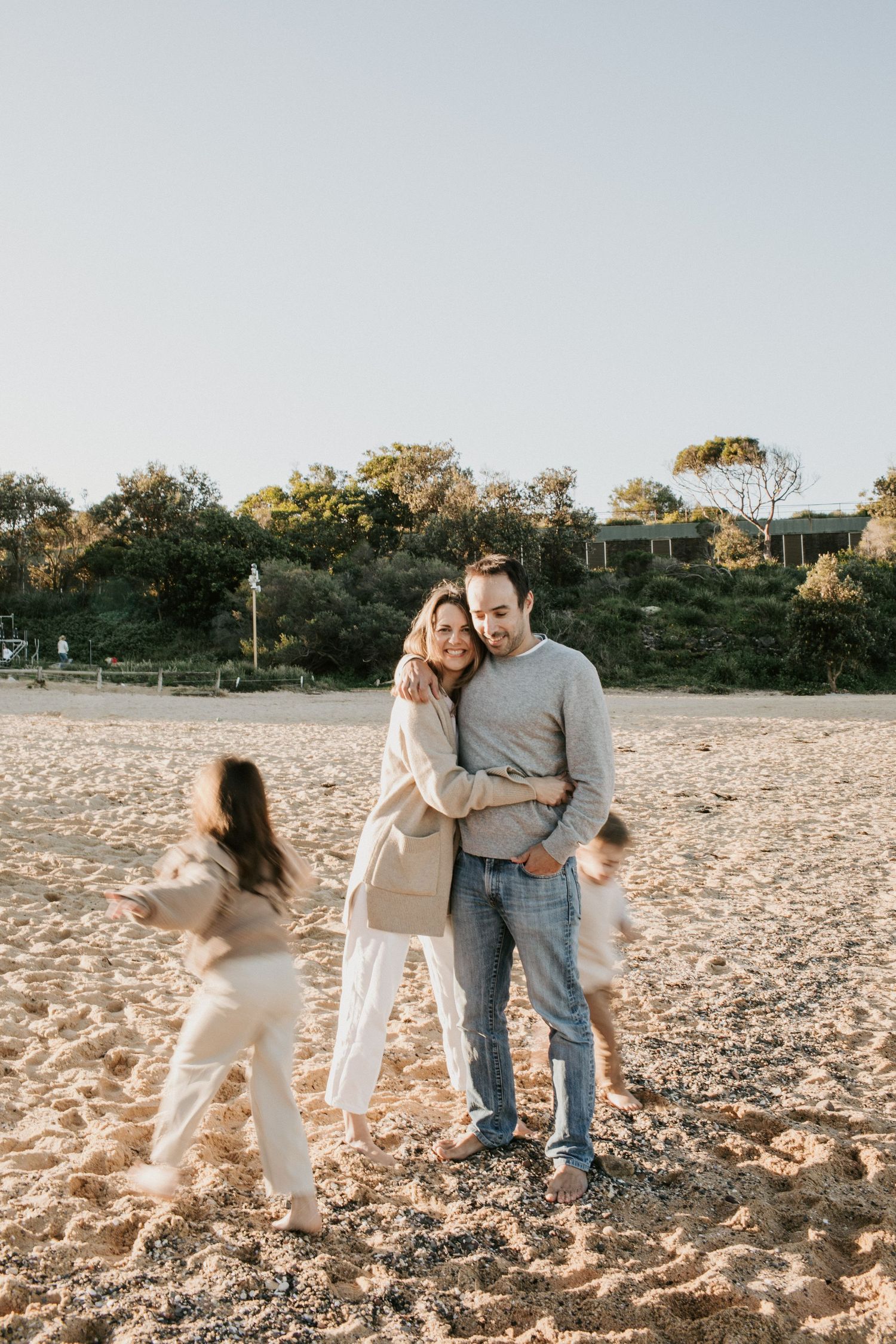 Couple embracing on a sandy beach while their children play around them.