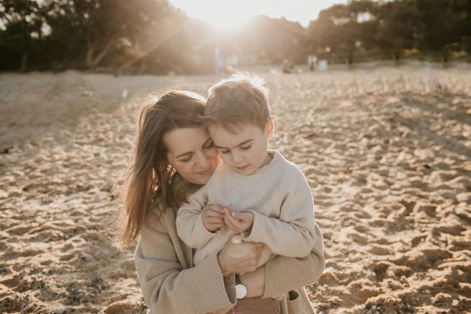 Mother tenderly hugging her young son on a sandy beach at sunset.
