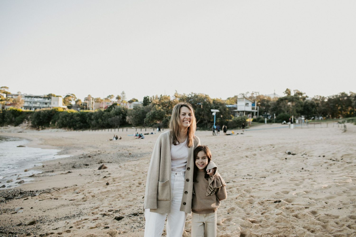 Smiling mother and daughter standing together on a sunny sandy beach with trees behind.