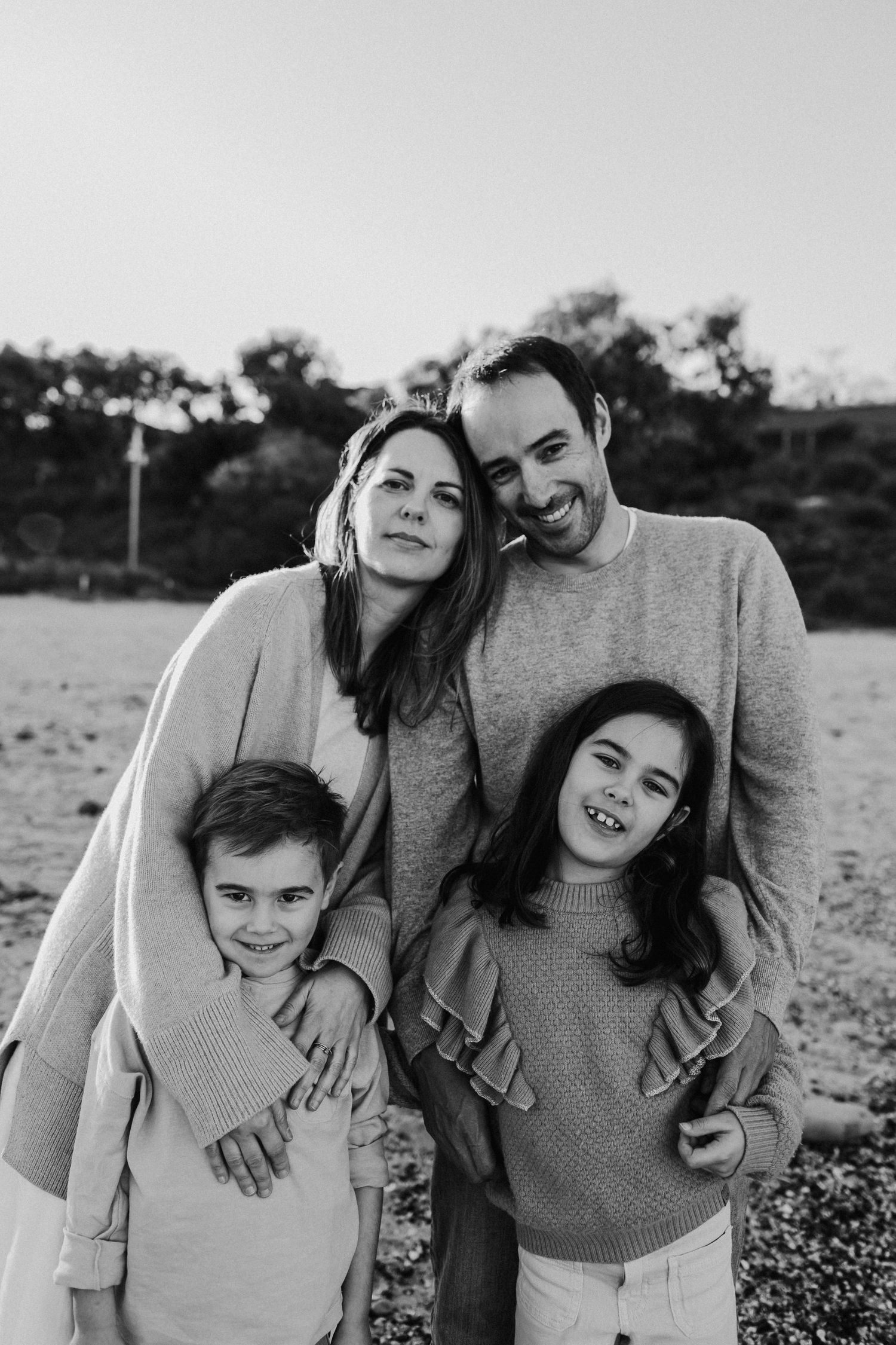 Black and white portrait of a happy family of four posing together on a beach at sunset.