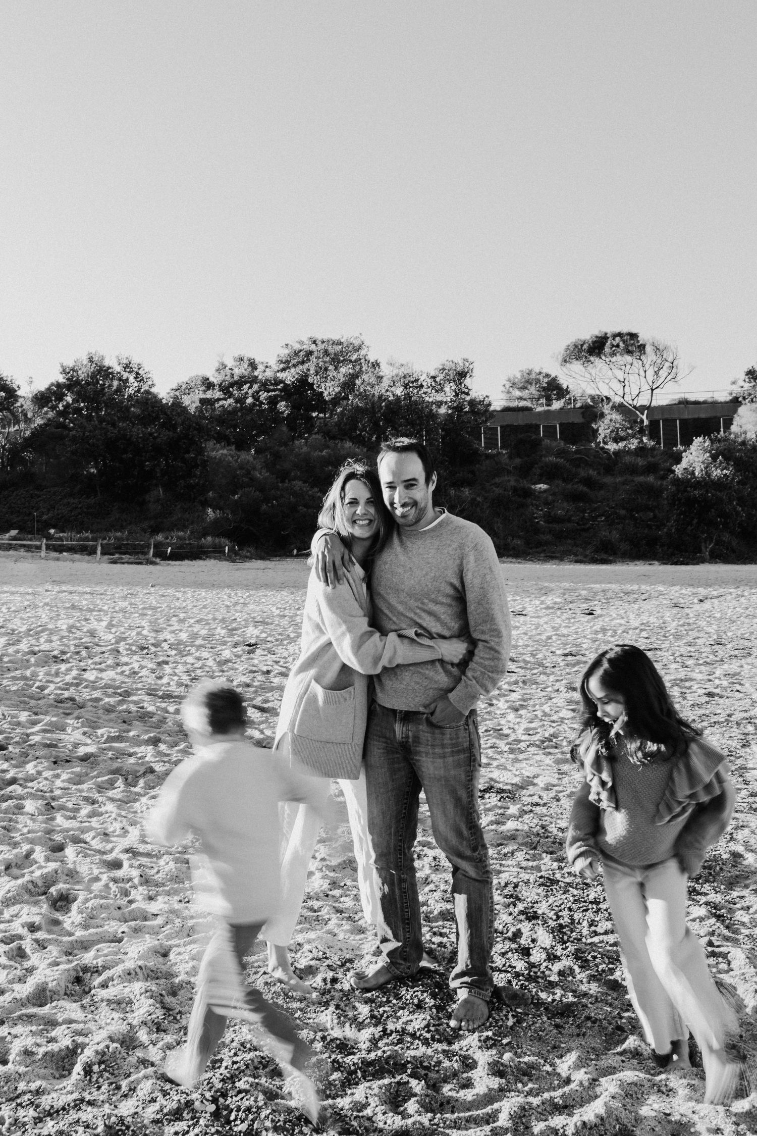 Black and white photo of a couple embracing on a sandy beach while two children play around them.