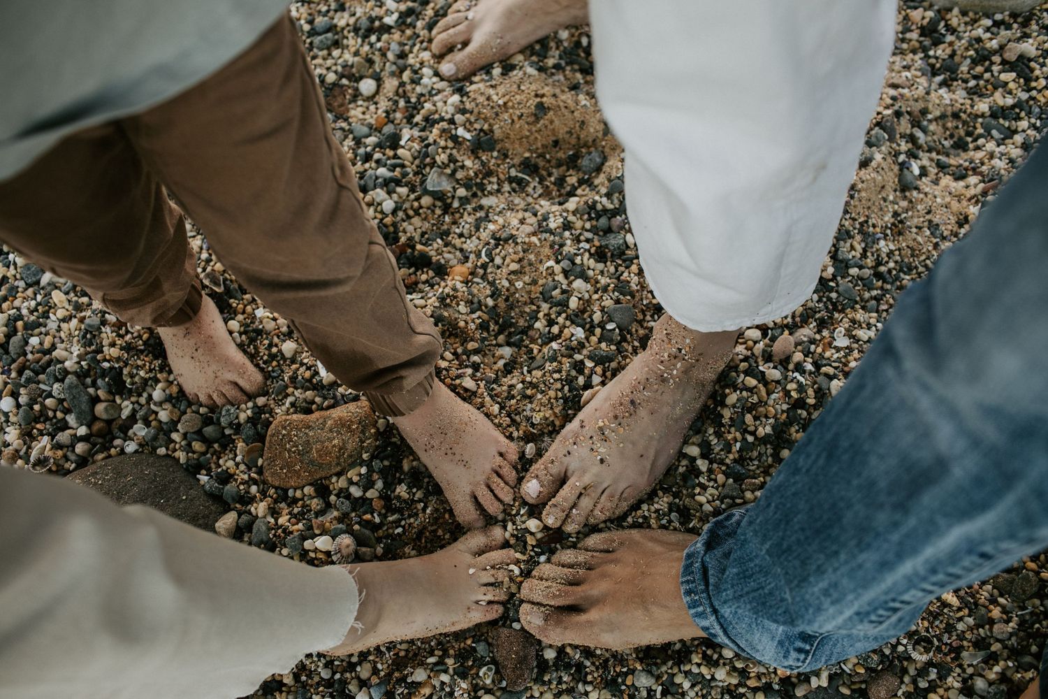 Four pairs of bare feet standing together on a rocky pebble beach from above.