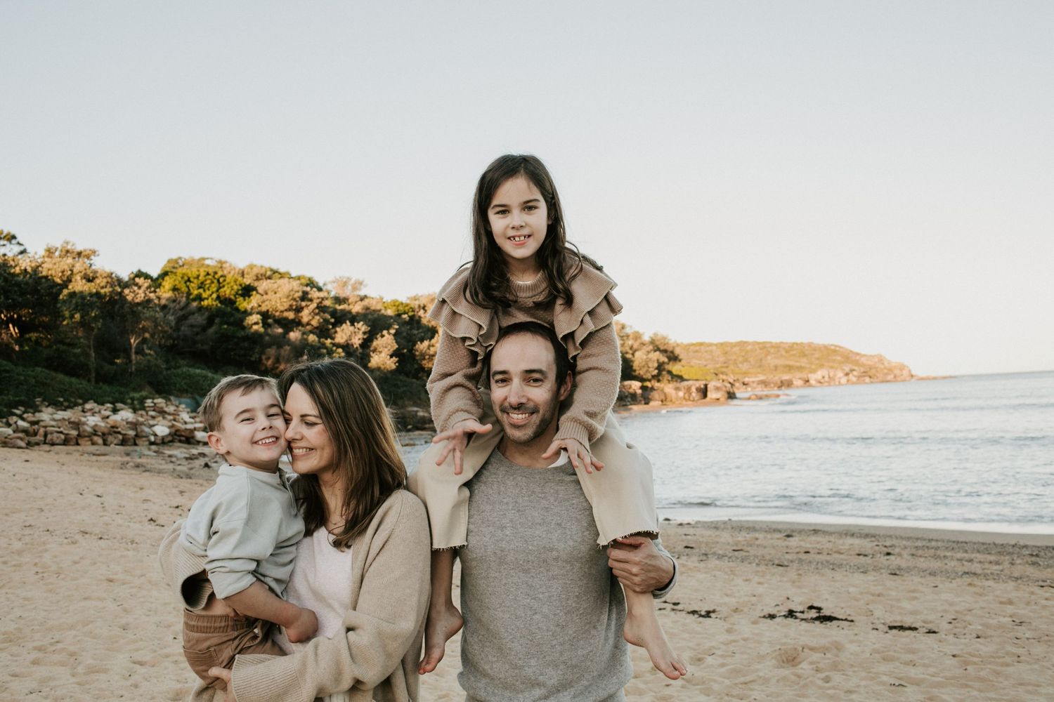 Happy family of four posing on a sandy beach at sunset with trees and water in the background.