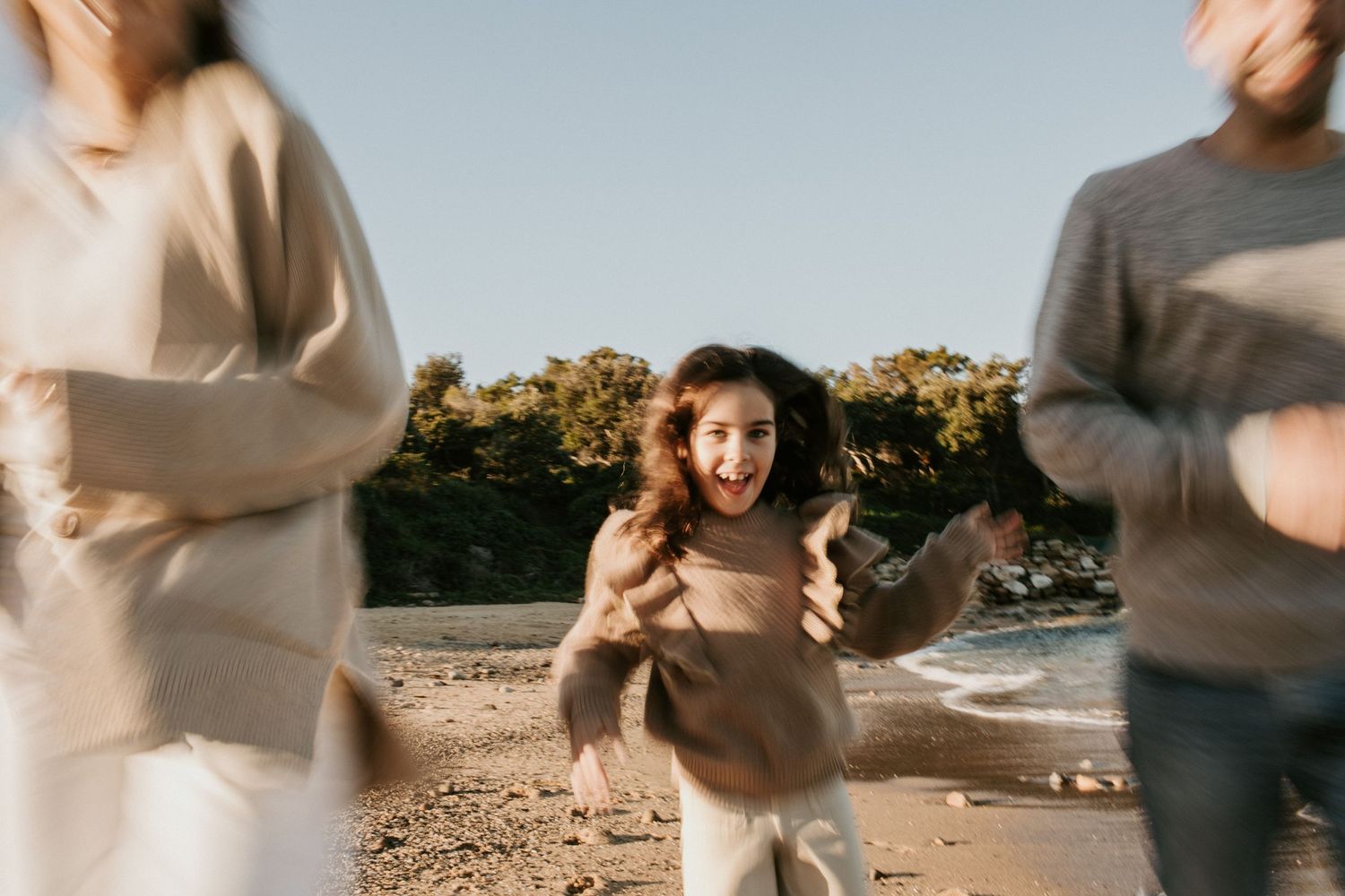 Excited young girl running toward camera on a sunny beach with parents blurred in foreground.