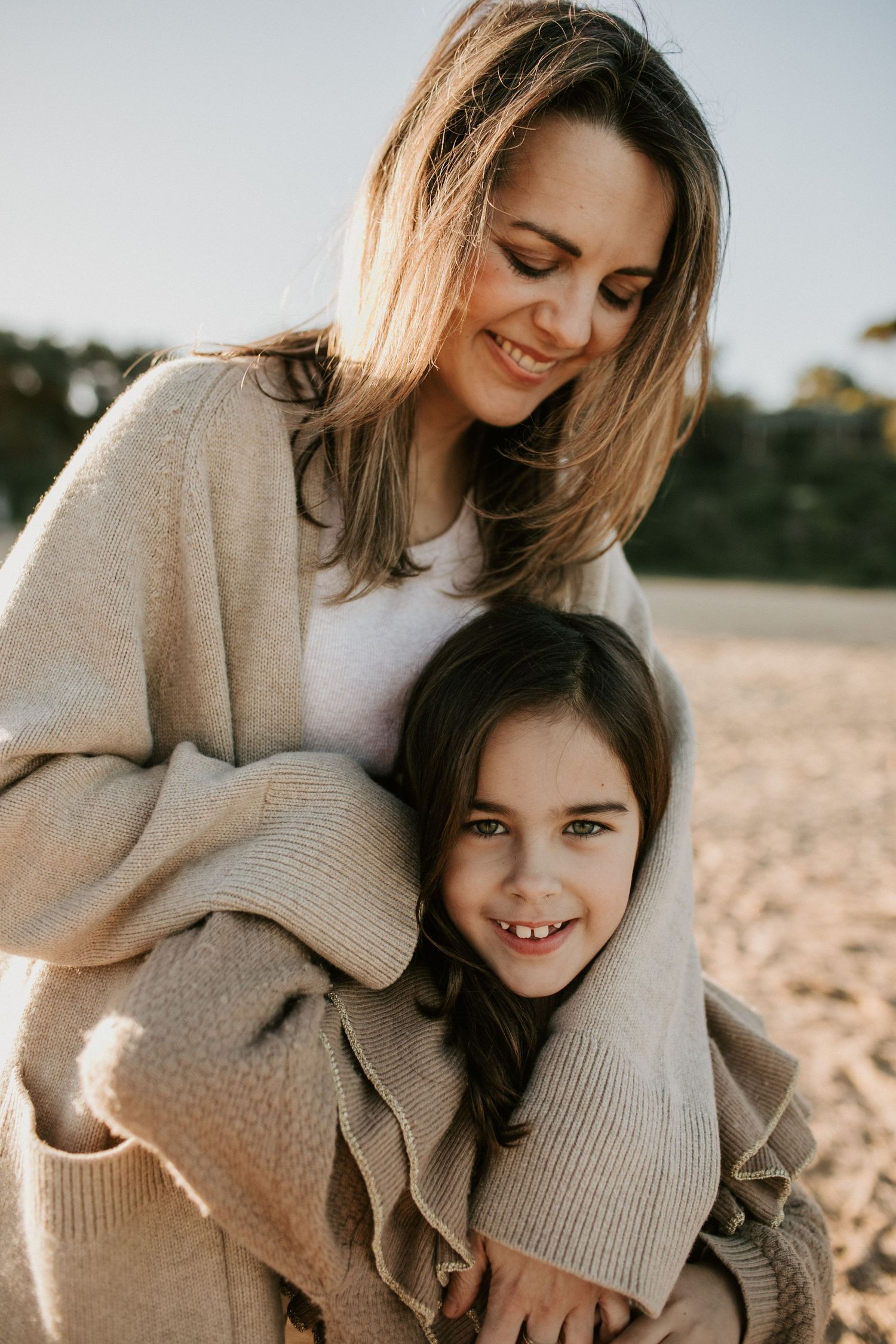 Smiling mother wrapping her cardigan around her happy young daughter on a sandy beach.