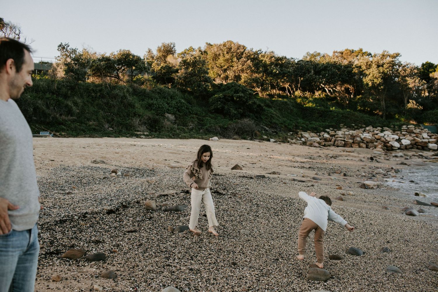 Father watches his two young children explore a rocky beach shore at golden hour.