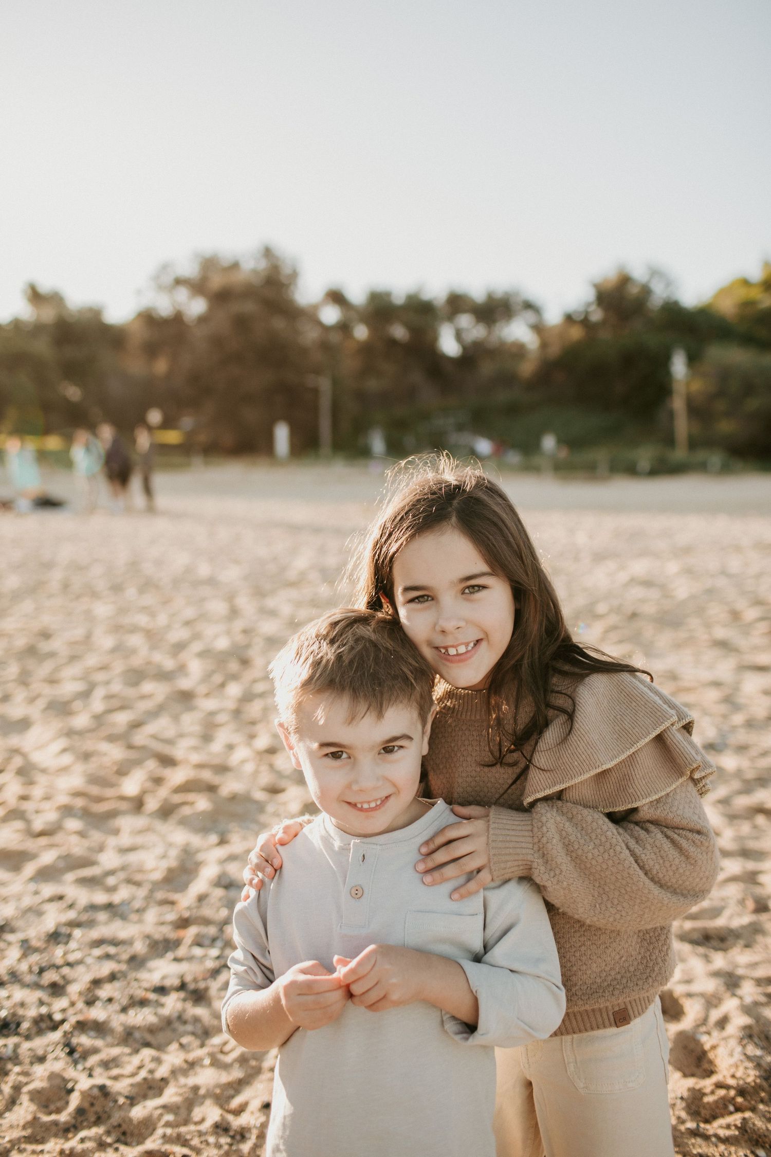 Two siblings hugging and smiling on a sunny beach, girl embracing younger boy from behind.