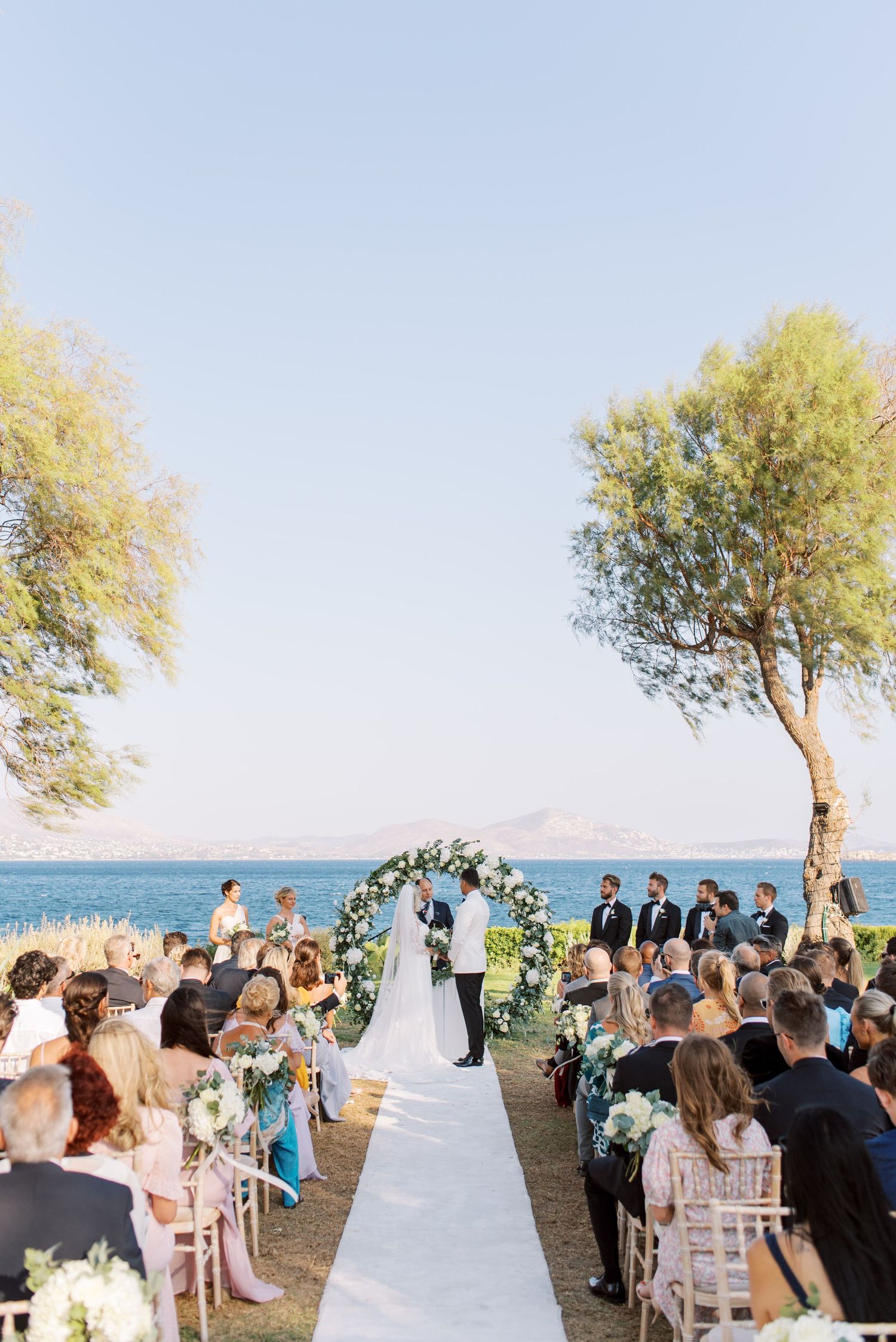 A seaside wedding ceremony with guests seated along a white aisle under trees overlooking the ocean.