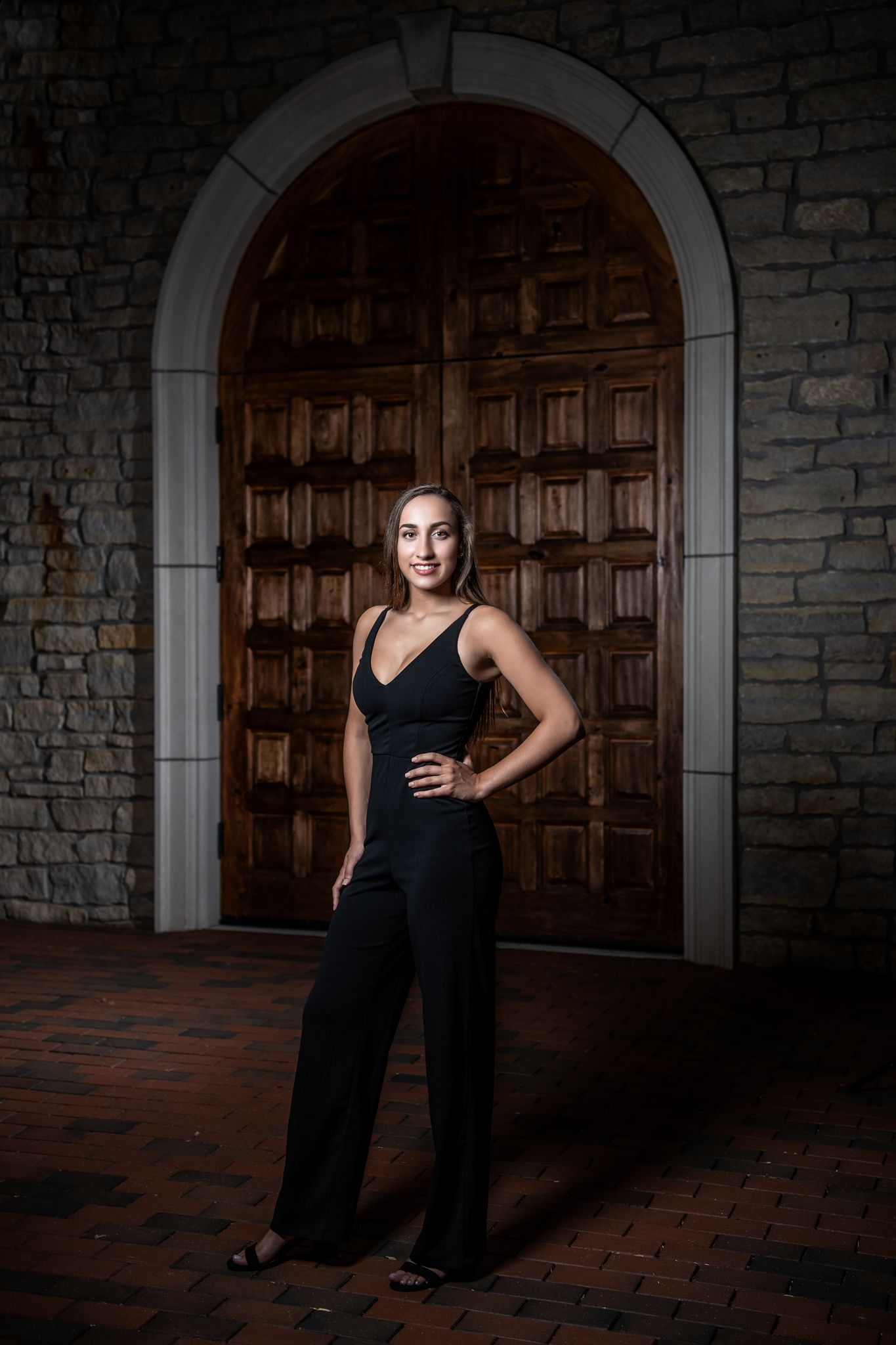 A dramatic portrait in front of a wooden arched door wearing an elegant black jumpsuit.