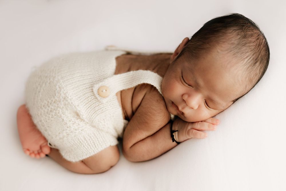Sleeping newborn wearing white knitted romper curled up on light background.