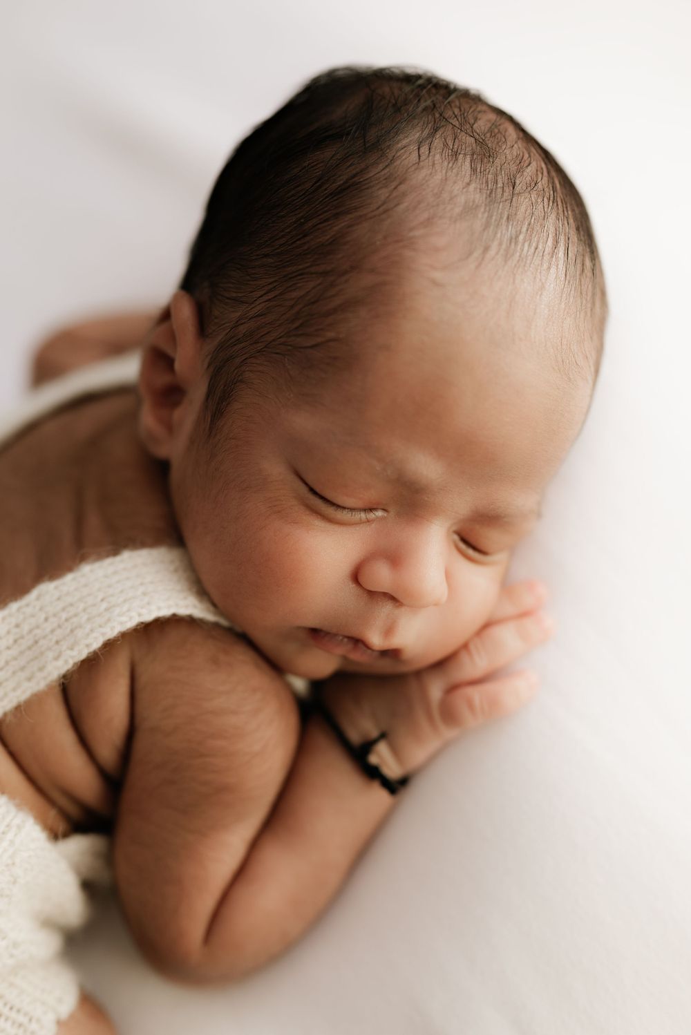Newborn baby in cream-colored outfit sleeping with hands tucked under chin.