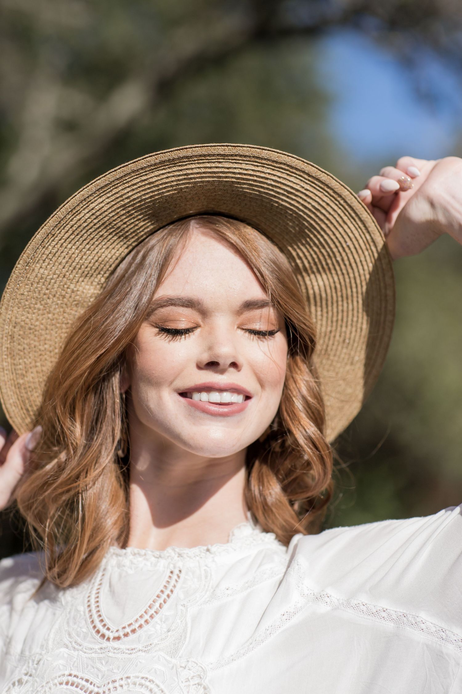 Smiling woman with eyes closed holding brim of straw hat wearing white dress in sunny garden.