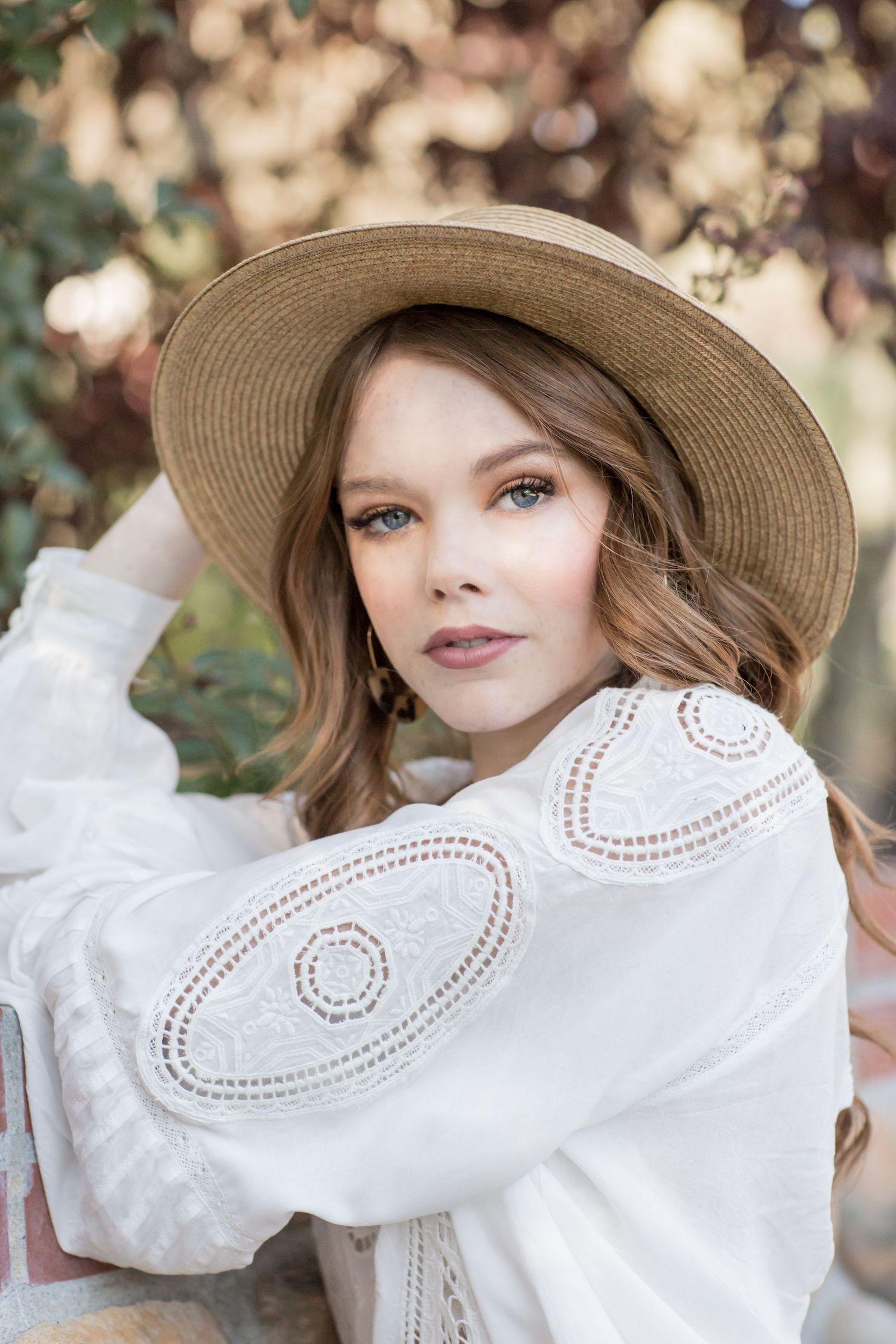 Close-up portrait of woman in straw hat and white lace top with embroidered details outdoors.