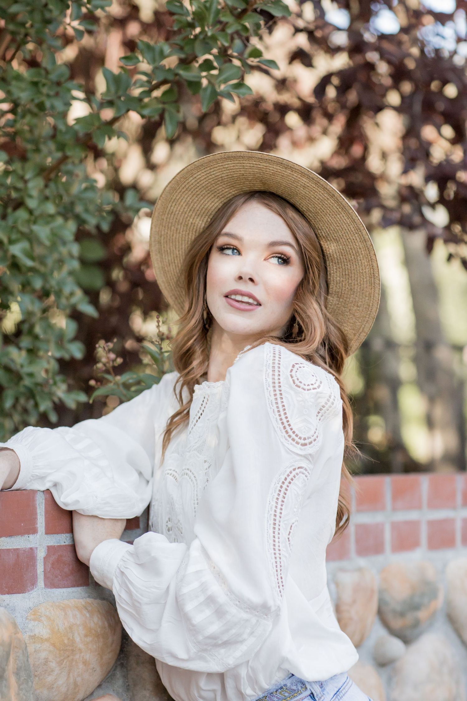 Young woman in white lace blouse and straw hat leaning on stone wall surrounded by greenery.