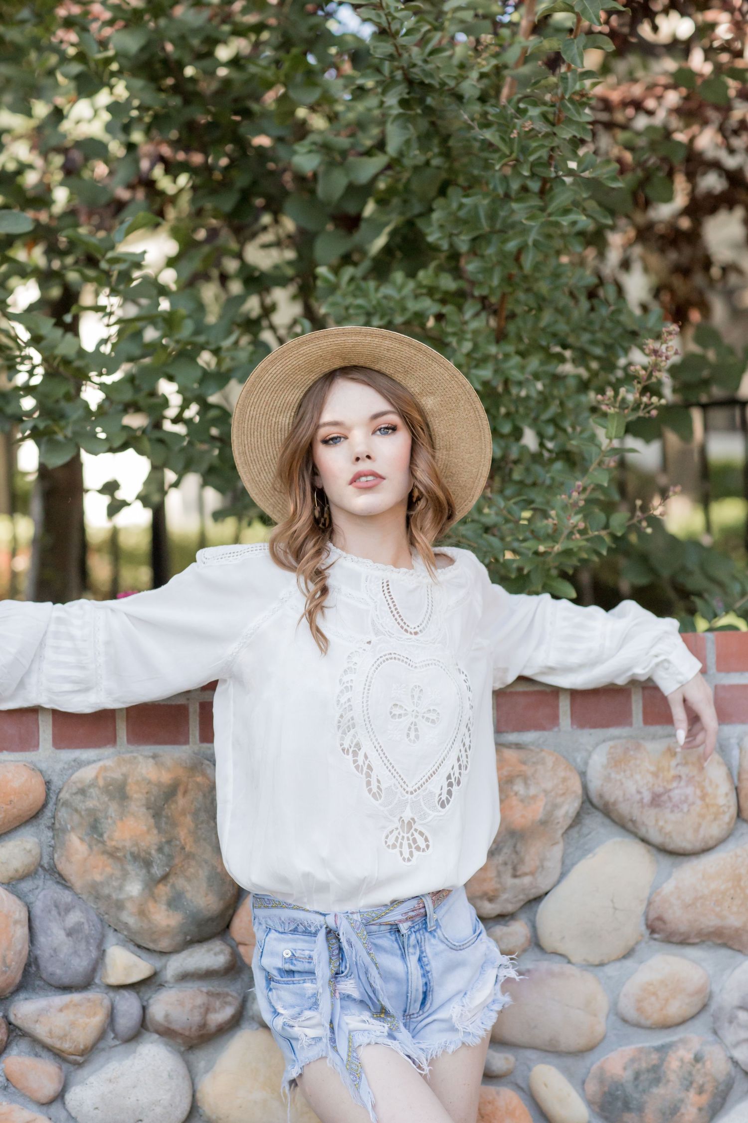 Young woman in embroidered white sweatshirt, denim shorts, and straw hat posing by stone wall.