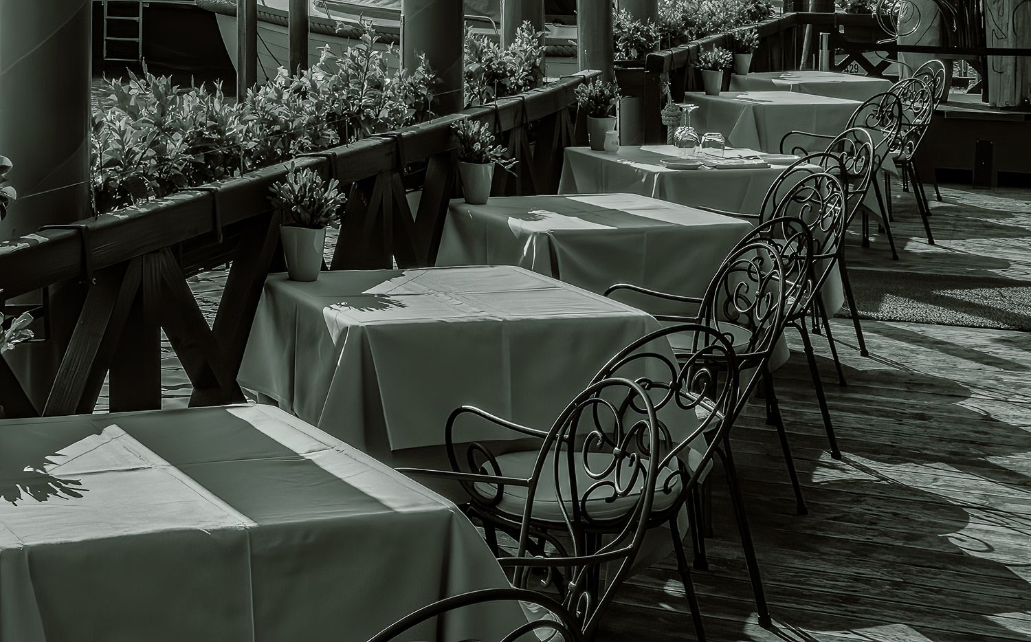 Black and white image of empty outdoor cafe tables with wrought iron chairs casting dramatic shadows.