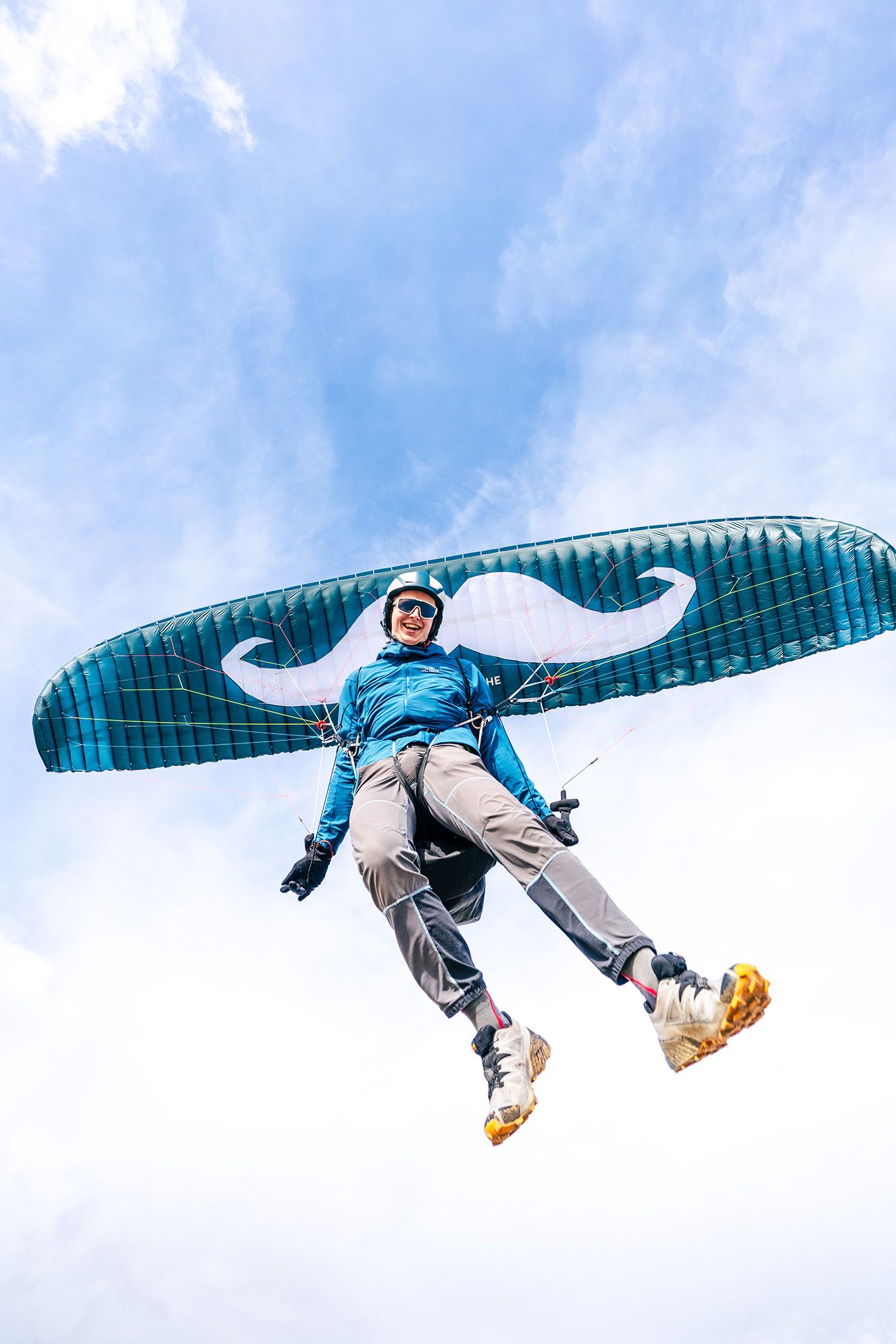 Paraglider soaring through blue sky with teal wing canopy overhead.