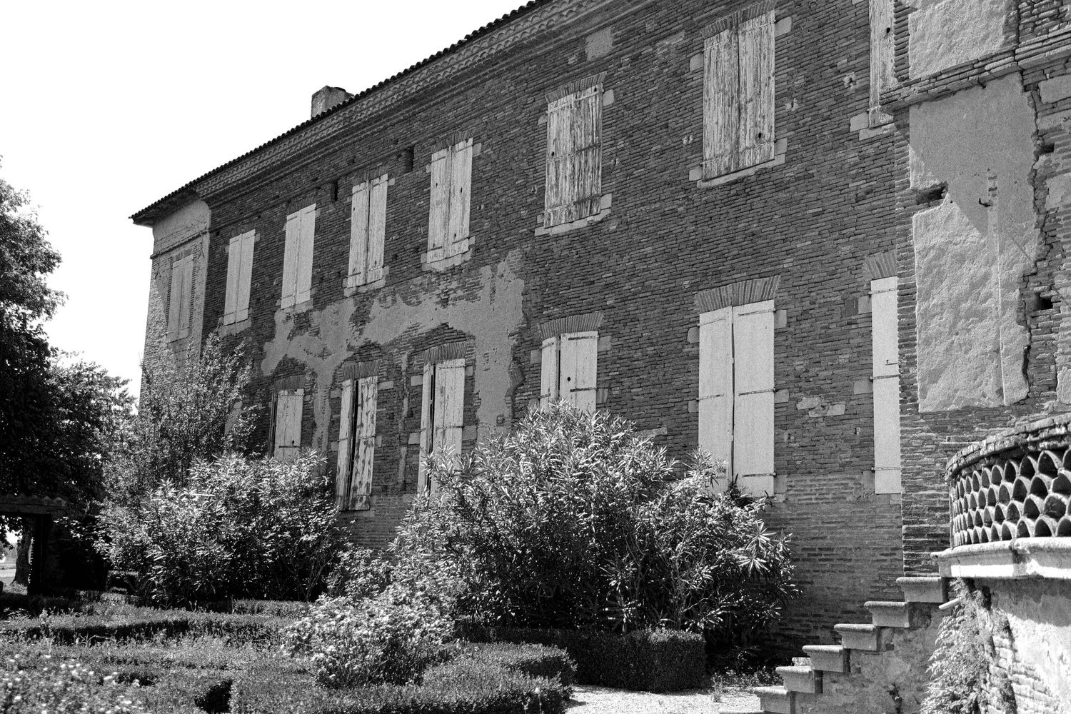 Historic brick mansion with white shuttered windows and overgrown garden shown in black and white photography.
