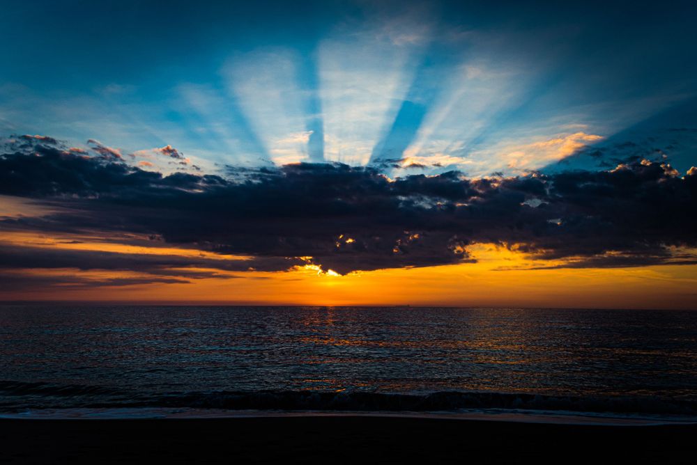 Dramatic sunset over ocean with sun rays bursting through dark clouds.