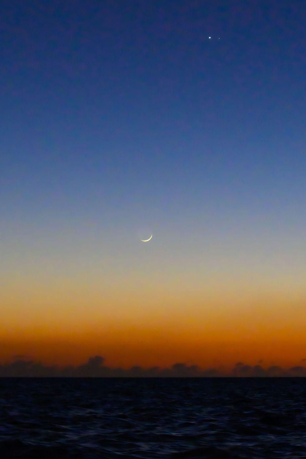 Crescent moon and bright star visible in gradient blue to orange twilight sky over ocean.