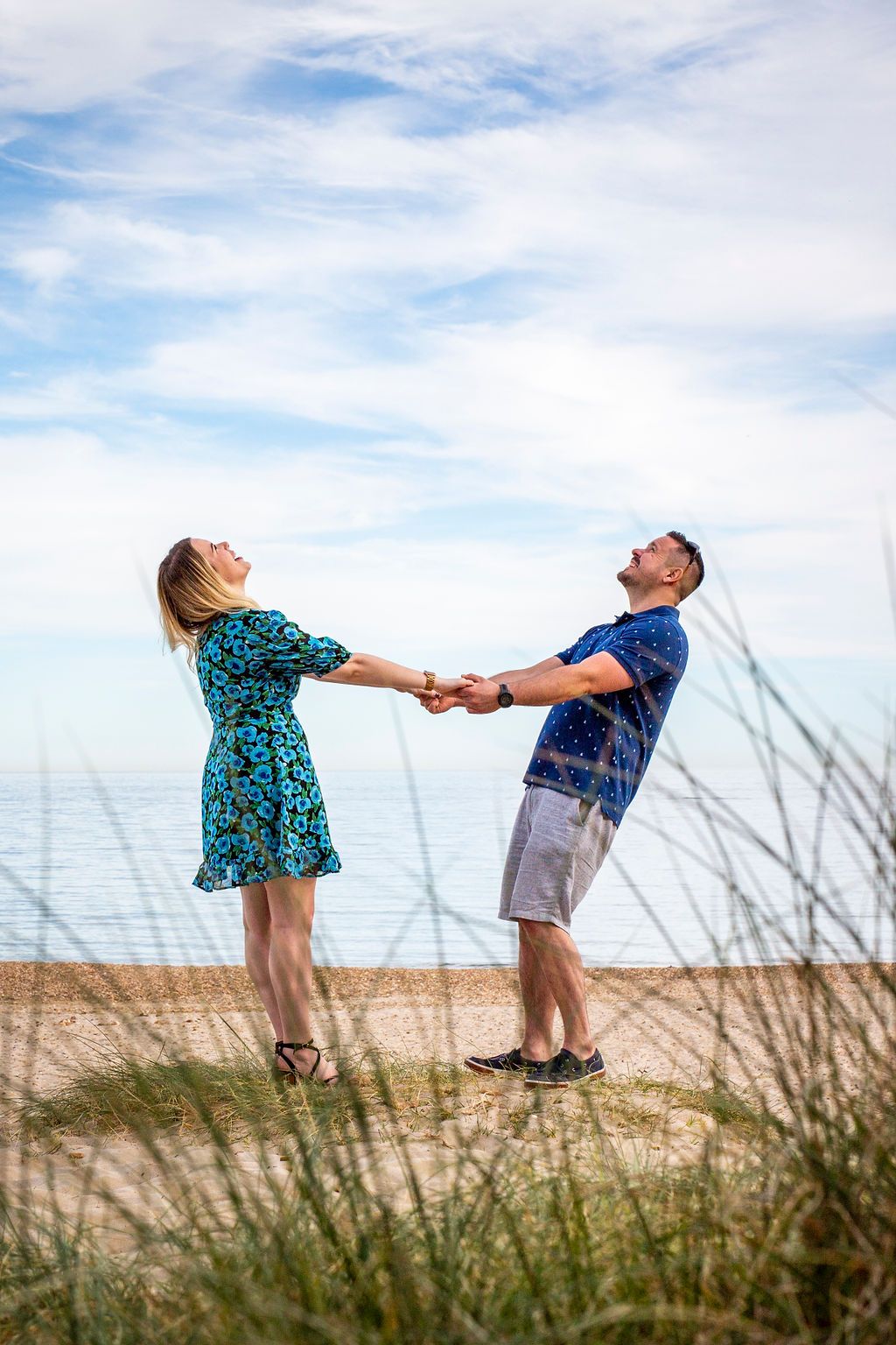 A playful couple holds hands and leans back on a beach with ocean views and blue skies.
