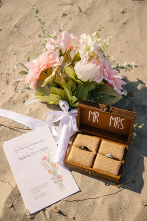 Wedding details with pink flowers, rings in wooden box marked Mr & Mrs, and invitation on sand.