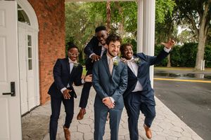 A group of groomsmen in suits posing playfully outside a brick building.