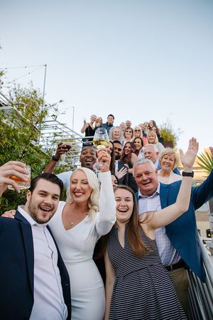 Joyful wedding party and guests celebrating with raised arms in an outdoor group photo.