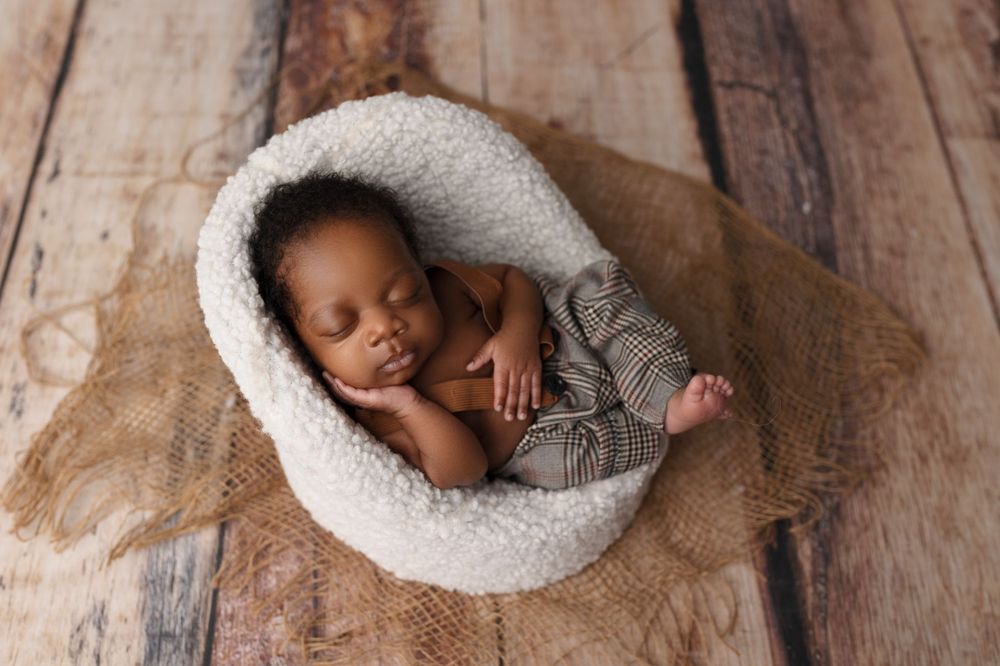 Sleeping baby in grey outfit curled up in cozy white round cushion on wooden backdrop.