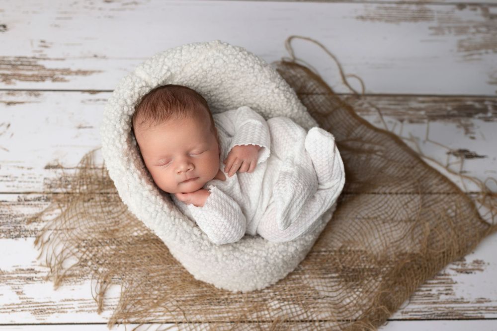Peaceful sleeping newborn in white outfit nestled in cream cushion on rustic wooden background.