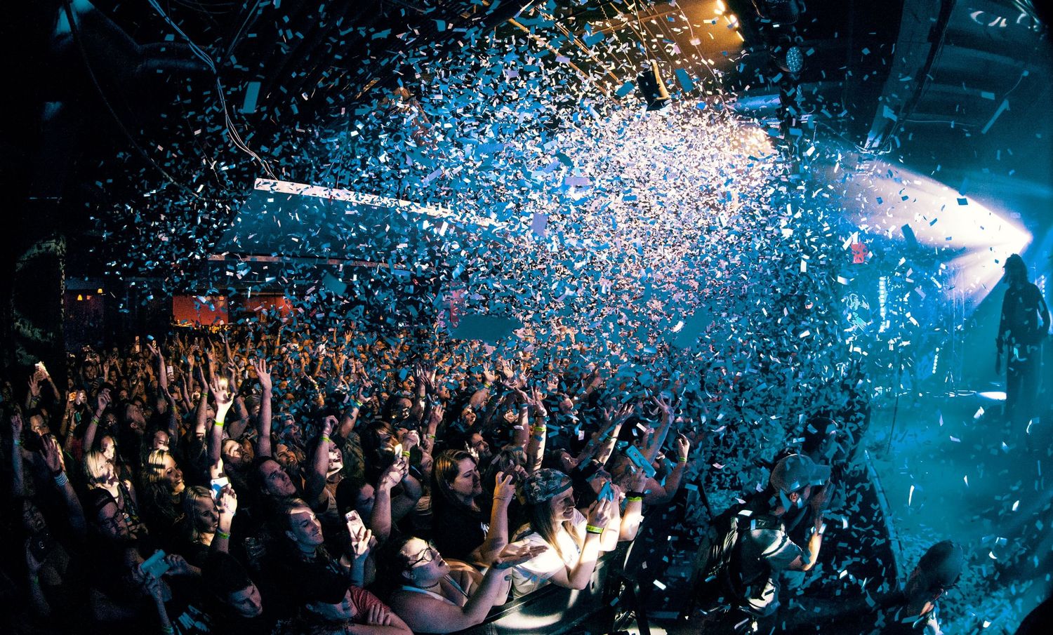 An energetic nightclub crowd beneath blue spotlights and glittering confetti.