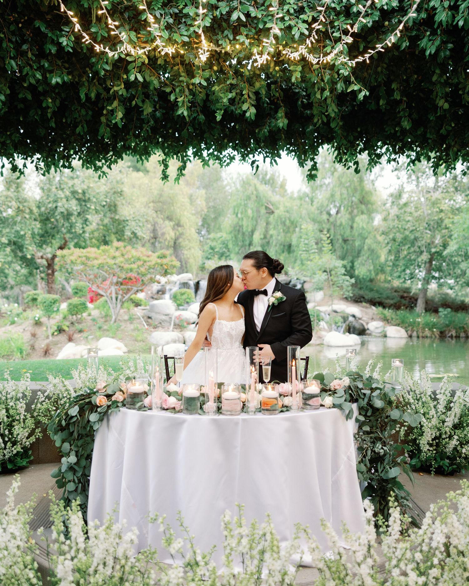 Bride and groom sitting at a romantically lit outdoor wedding reception table draped in greenery and fairy lights.
