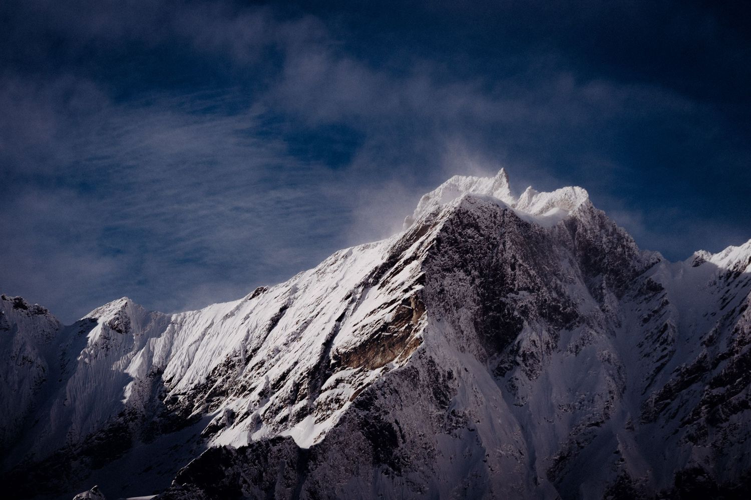 Snow-covered mountain peak against a dramatic dark blue sky with wispy clouds.