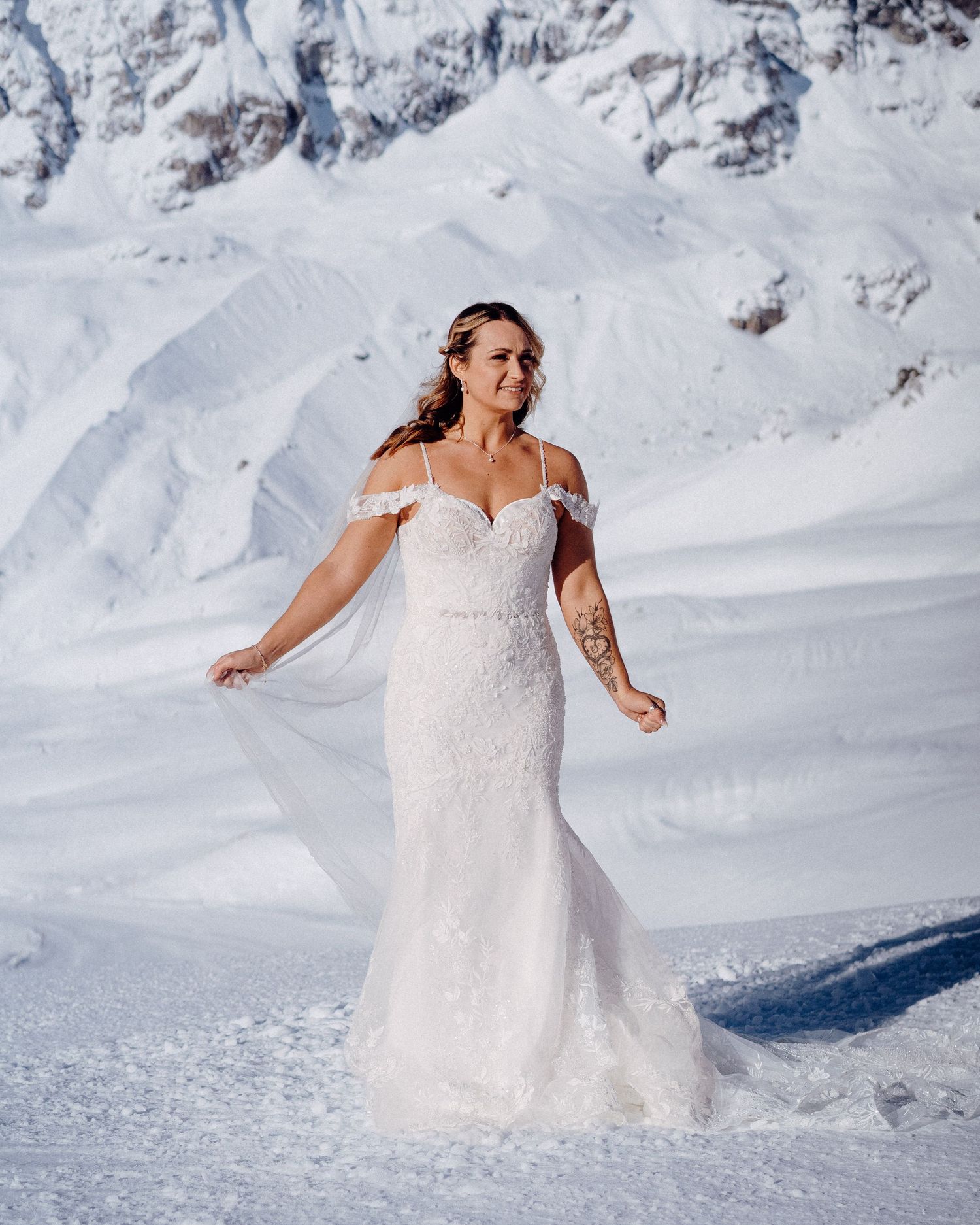 Bride in a white lace dress poses in a snowy mountain landscape.
