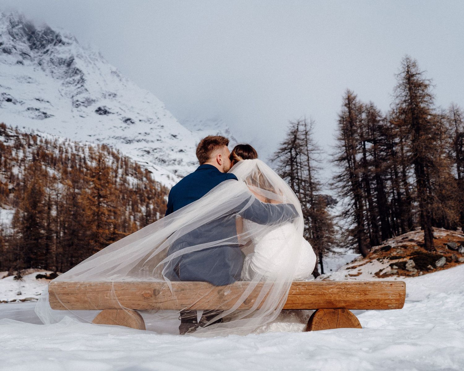 Couple sharing an intimate moment on a wooden bench surrounded by snow and mountains.