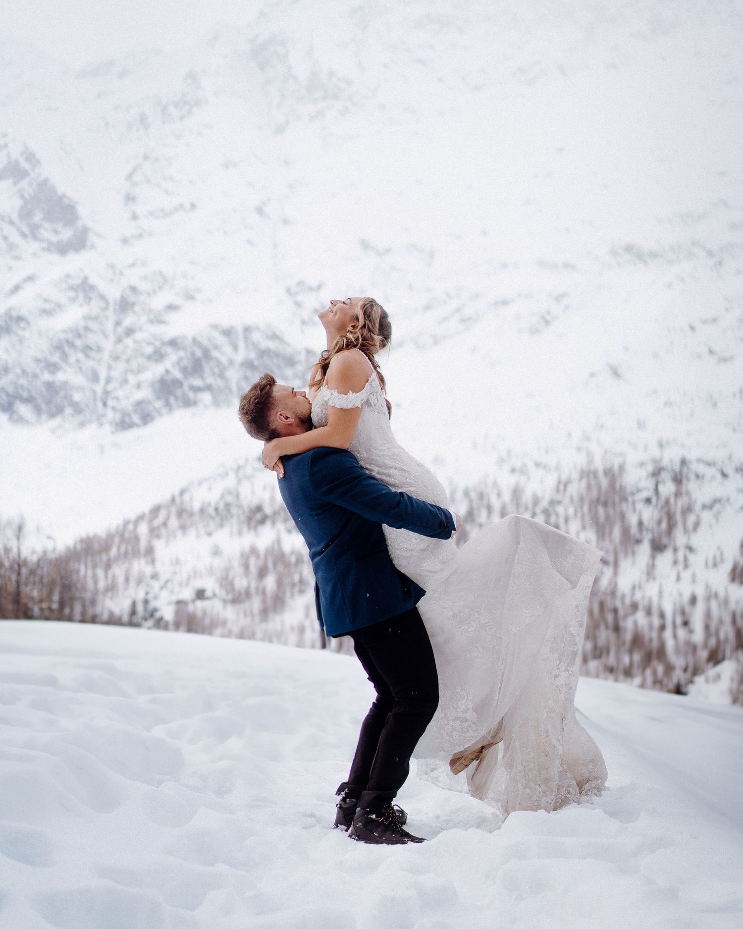 Wedding couple embraces joyfully in a snowy mountain landscape during their winter celebration.