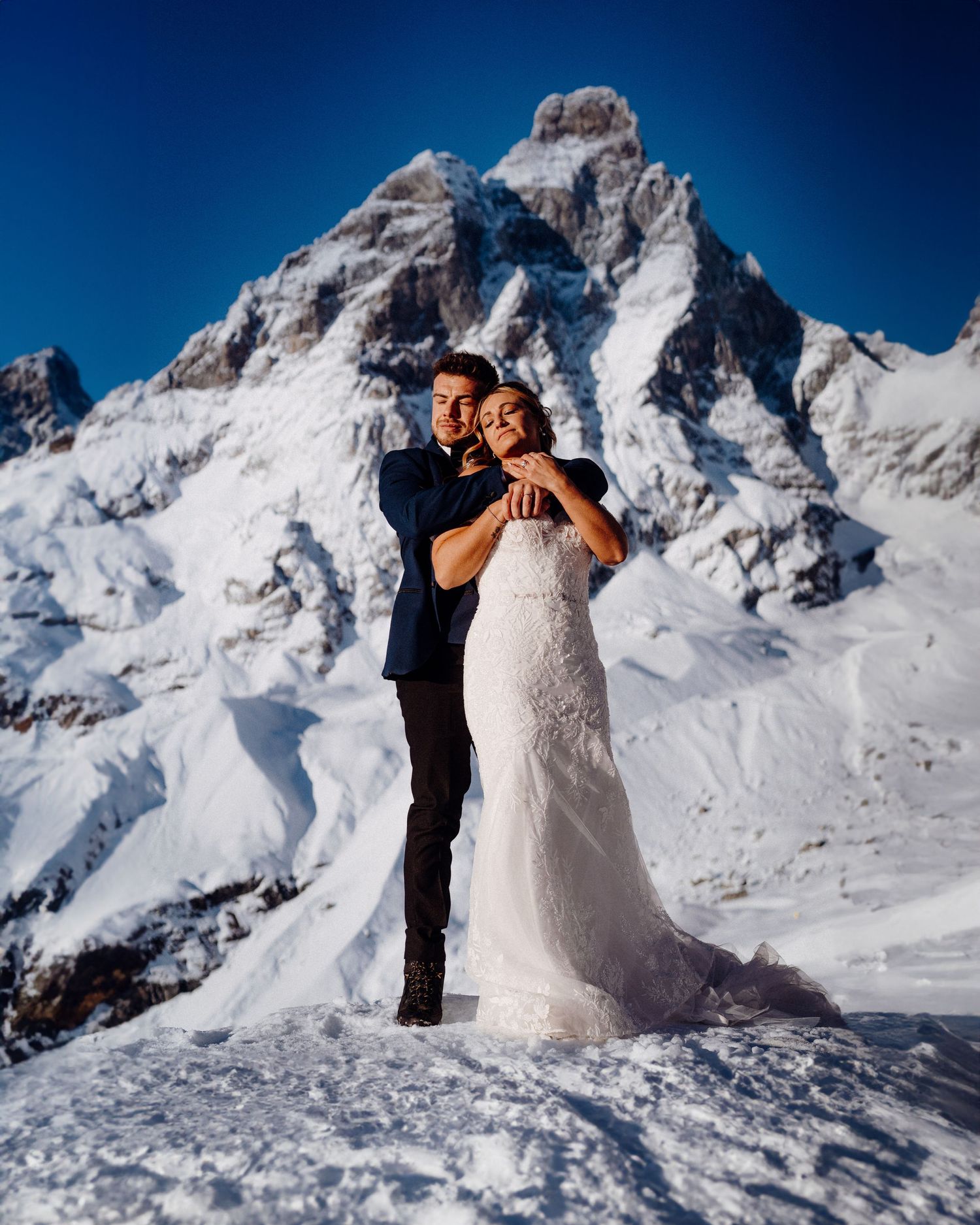 Newlyweds embrace in front of snow-capped mountains on a bright winter day.