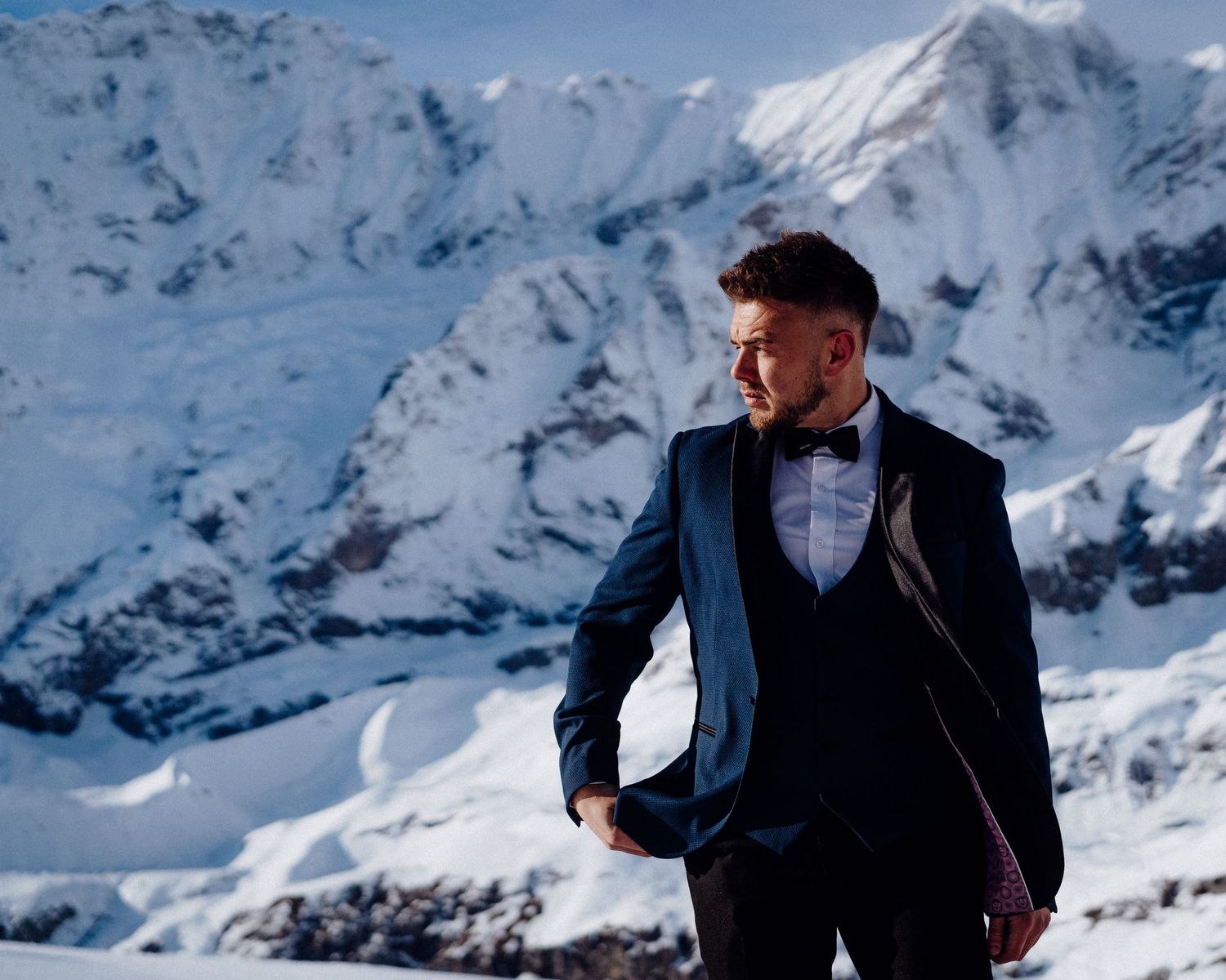 A groom in a navy suit poses against a stunning snowy mountain backdrop.