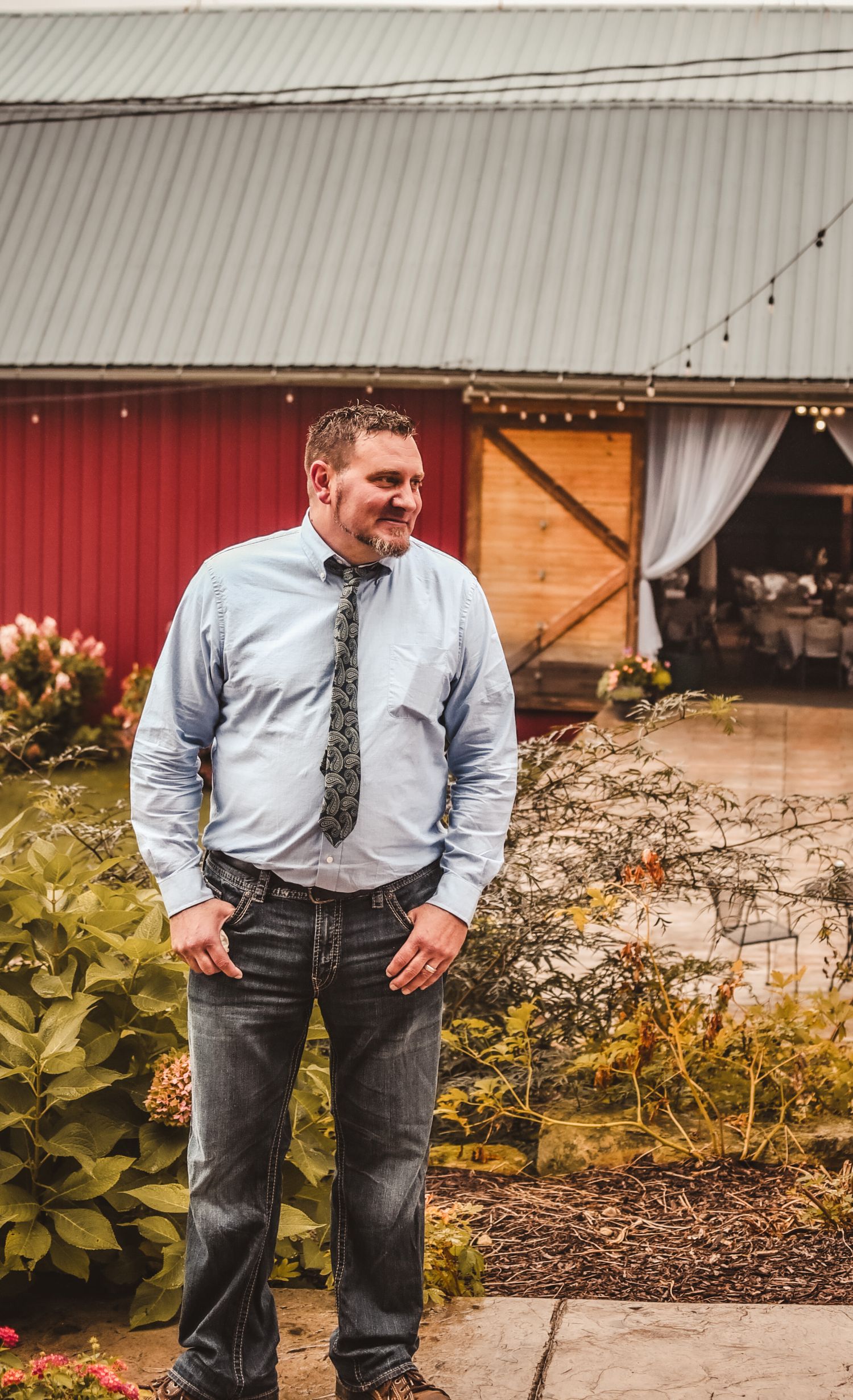Man in light blue shirt and tie standing by red barn with string lights.