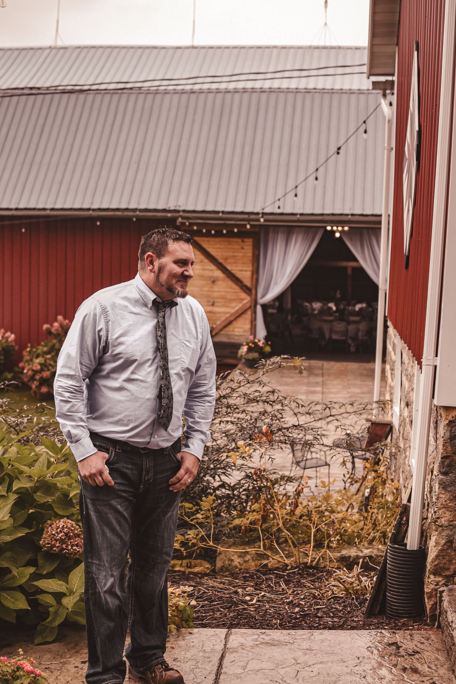 A person in a light button-down shirt stands thoughtfully outside a red barn with string lights.