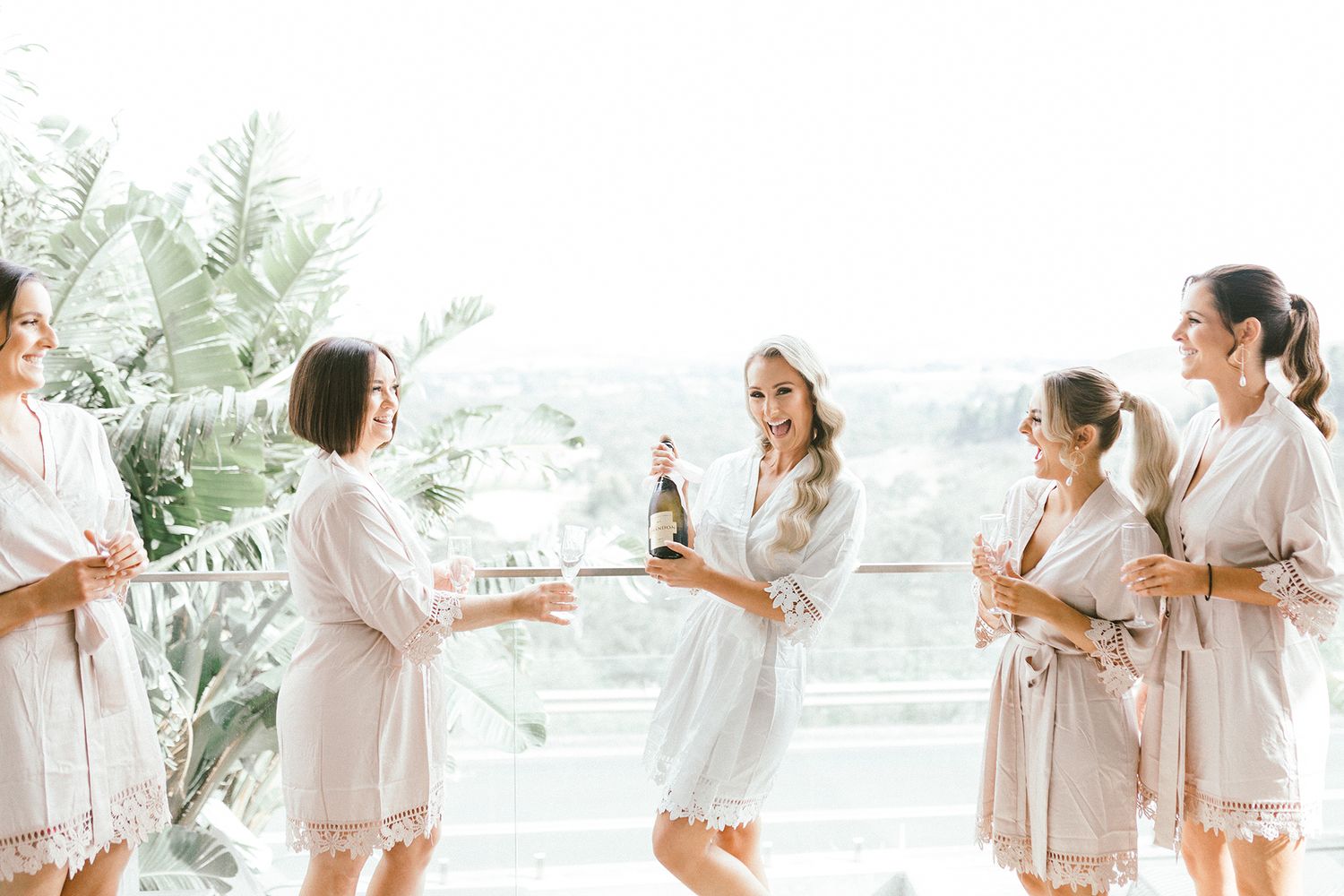 Wedding party in matching robes celebrating with champagne in bright sunlit room.