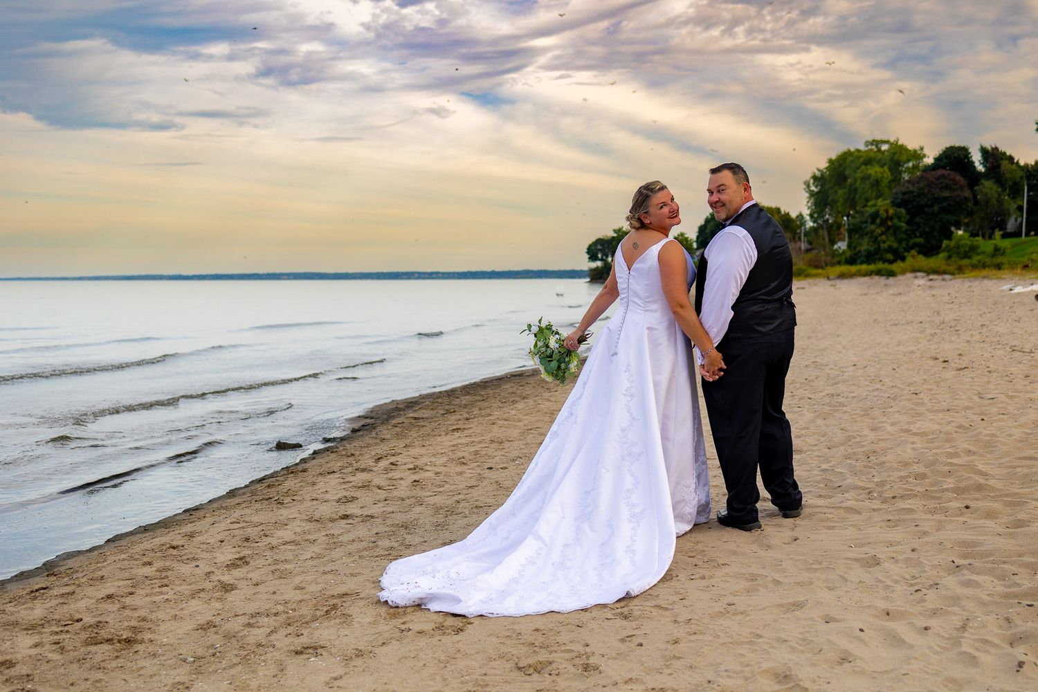 A wedding couple poses on a beach at sunset with ocean waves in the background.