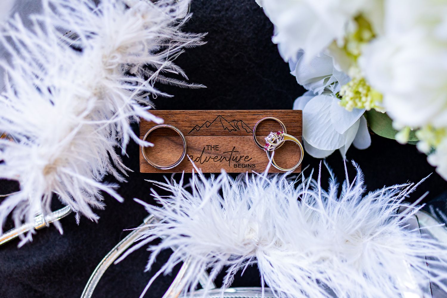 Wedding rings displayed on a wooden holder against white feathers and floral decorations.