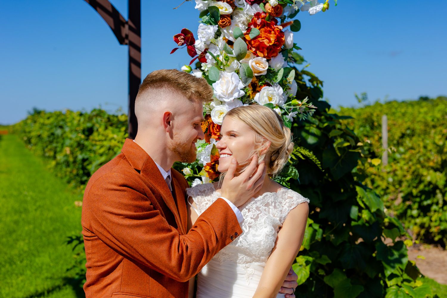 Newlyweds embrace under a floral arch in a vineyard on their wedding day.