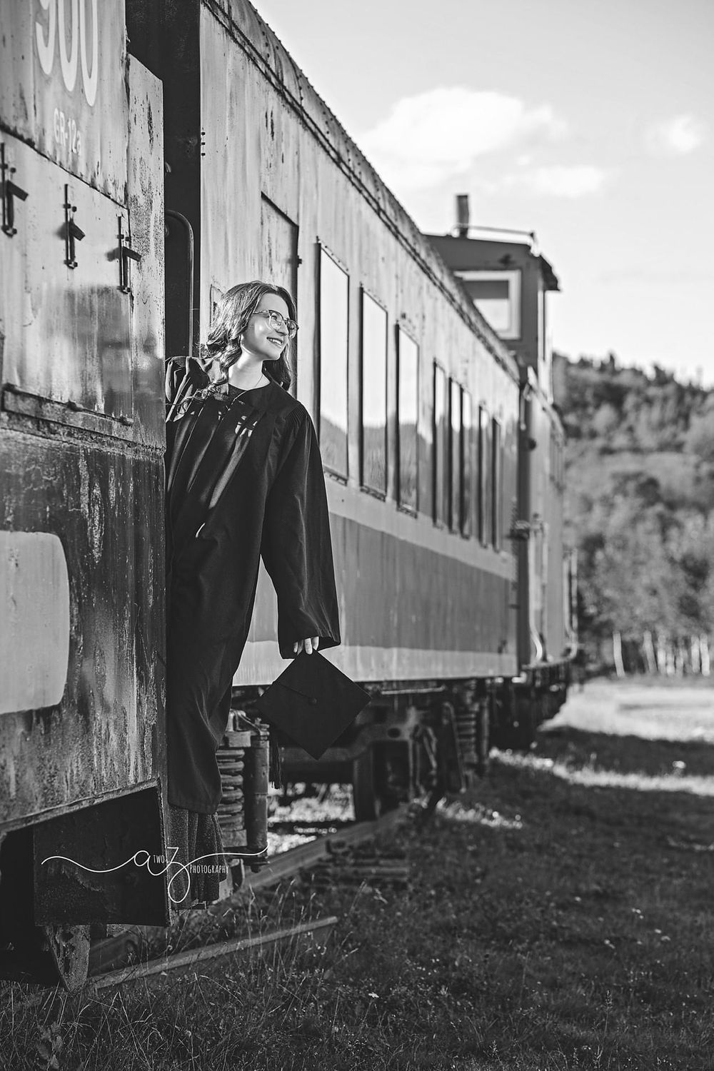Graduate in cap and gown posing on vintage train car in black and white photograph.
